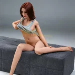 Nuvielle Sex Doll 155 cm IronTech posée sur un banc, enlevant un haut gris, cheveux bruns.