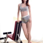 Ondine Sex Doll 168 cm IronTech en tenue de sport grise, posée près d'un banc de musculation.