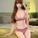 Ruby Sex Doll 163 cm IronTech assise sur un lit, lingerie rose, cheveux longs bruns.