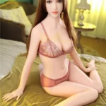 Ruby Sex Doll 163 cm IronTech Doll TPE assise sur un lit, lingerie rose, cheveux longs.