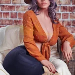 Velena Sex Doll 156 cm en tenue western, chemisier orange, chapeau et bottes, posant assise.