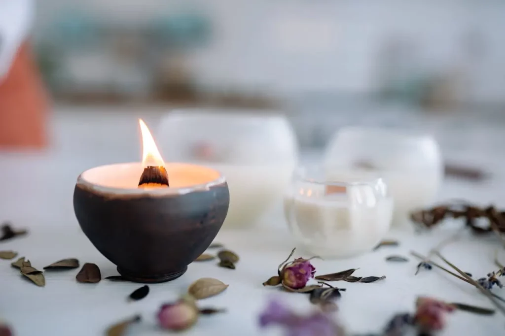 Bougie allumée dans un récipient noir entourée de fleurs séchées et d'autres bougies blanches en arrière-plan, créant une atmosphère intime et relaxante