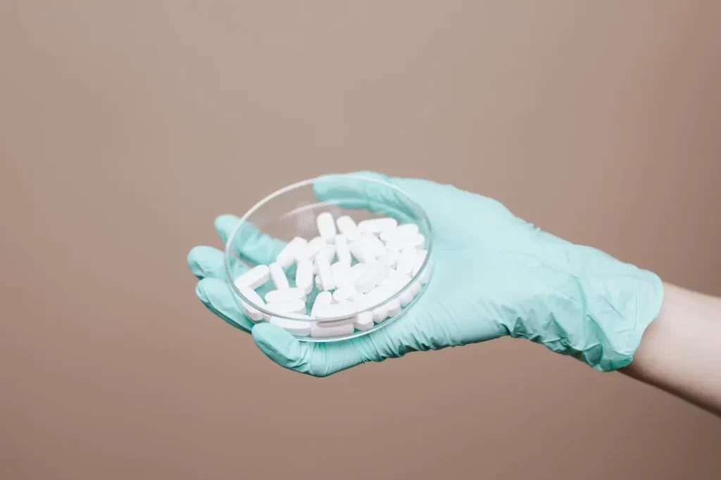 Main gantée de scientifique distribuant des comprimés blancs dans un récipient transparent, symbolisant les solutions médicamenteuses pour l’éjaculation précoce.