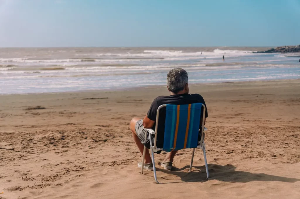 Homme senior vu de dos, assis nonchalamment sur une chaise de plage face à l’océan, incarnant le calme et le bien-être retrouvés grâce au soutien discret d’une Real Doll thérapeutique