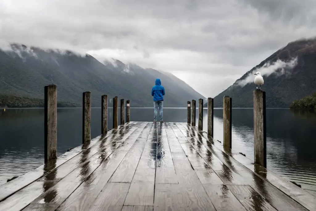 Homme seul de dos sur un ponton en bois face à un lac et des montagnes par temps gris, illustrant la solitude après une rupture amoureuse.